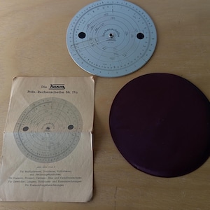 Może przedstawiać: Zestaw zabytkowych suwaków, zawierający okrągły suwak, brązowe okrągłe etui i papierową instrukcję. Suwak jest jasnoniebieski z czarnymi oznaczeniami. Instrukcja jest w kolorze beżowym z czarnym tekstem, w tym "Die Hansa".