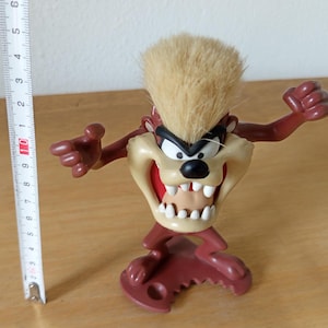 Puede incluir: Una figura de Taz, un personaje de dibujos animados, de pie con los brazos extendidos. La figura mide aproximadamente 10 cm de altura. Tiene una cara de color beige, una boca roja y un cuerpo marrón. La figura tiene un diseño de pelo espinoso de color marrón claro en la parte superior de la cabeza.