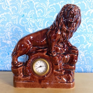 Puede incluir: Una figura de león de cerámica marrón con un reloj integrado. El león está posado sobre una base rocosa, con la esfera del reloj incrustada en la base. El reloj tiene un bisel dorado y una esfera blanca con números y agujas negras.