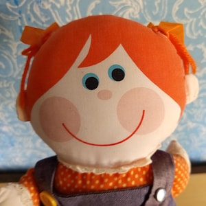 Puede incluir: Una muñeca vintage con cabello naranja en coletas, ojos azules y mejillas rosadas. La muñeca lleva un vestido con peto morado con una camisa naranja con lunares debajo. Un juguete clásico con una cara sonriente.