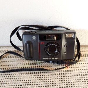Può includere: Una fotocamera a pellicola compatta Minolta Riva Zoom 70c grigio scuro con tracolla nera. La parte anteriore presenta un obiettivo Minolta con zoom 35-70mm, un flash, un mirino e il testo Minolta, E5 e Riva Zoom 70c stampato sul corpo macchina.