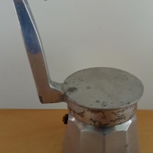 Puede incluir: Una cafetera italiana vintage de color plateado. La cafetera tiene una base hexagonal y una parte superior redonda con un asa. El asa tiene un diseño curvo y angular. La cafetera está hecha de metal.