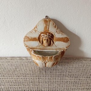 May include: A wall-mounted holy water font featuring a central relief sculpture of the face of Jesus wearing a crown of thorns. The piece is crafted from a light-colored material with brown antiqued detailing and includes a small basin at the bottom.