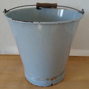 May include: A light blue enamel bucket with a metal handle and a wooden grip. The bucket has a slightly tapered shape and shows signs of wear around the rim and base, suggesting age and use. The handle is attached to the bucket with metal brackets.