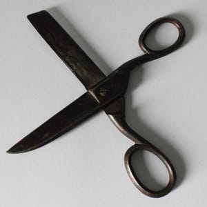 Puede incluir: Primer plano de un par de tijeras antiguas. Las tijeras son de metal oscuro con un aspecto ligeramente desgastado. Las hojas están cruzadas y los mangos tienen orificios ovalados para los dedos.