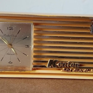 Puede incluir: Radio reloj Kenton de estado sólido vintage. La esfera del reloj es plateada con números y manecillas negras. La radio tiene un exterior dorado con listones horizontales y el logotipo de Kenton.