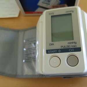 Puede incluir: Un tensi&oacute;metro Omron blanco y gris con pantalla digital. El dispositivo tiene un manguito y dos botones con la etiqueta "START" y "O/I". La pantalla muestra lecturas en mmHg y pulsaciones/min.