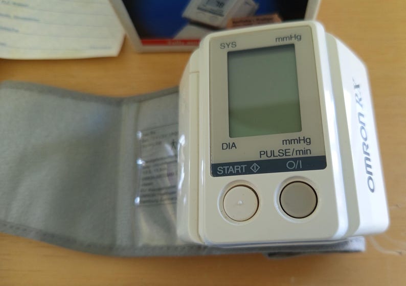 Puede incluir: Un tensi&oacute;metro Omron blanco y gris con pantalla digital. El dispositivo tiene un manguito y dos botones con la etiqueta "START" y "O/I". La pantalla muestra lecturas en mmHg y pulsaciones/min.