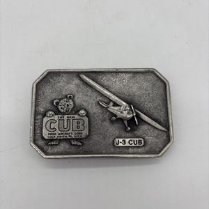 Könnte beinhalten: Eine rechteckige Gürtelschnalle aus silberfarbenem Metall mit einem geprägten Design eines kleinen Flugzeugs und eines Bären, der ein Schild mit der Aufschrift THE NEW CUB PIPER AIRCRAFT CORP. LOCK HAVEN, PA. U.S.A. hält. Der Text J-3 CUB ist ebenfalls eingeprägt.
