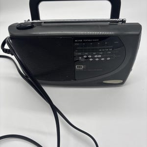 Peut inclure: Une radio portable grise foncée avec une poignée et une antenne noires. La radio a un haut-parleur et un cadran de réglage avec le texte "AE 2145 PORTABLE RADIO". Un cordon d'alimentation noir est également visible.
