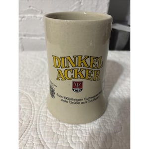 May include: A beige ceramic beer stein with the words "DINKEL ACKER" in yellow lettering. Below the text is a red and white logo. The stein has a cylindrical shape and a slightly flared base. Additional text is printed below the logo.