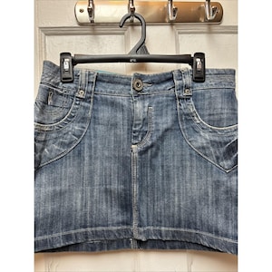 May include: A denim mini skirt in a medium wash blue. The skirt features a classic five-pocket design with decorative stitching and a button closure. It is hanging on a black hanger against a white door.