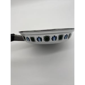 May include: White enamel frying pan with a black handle and a decorative floral pattern in green and blue. The pan has a silver rim and is angled to show the interior design.