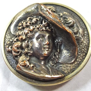 May include: A circular metal button featuring a high-relief, silver-toned portrait of a person wearing a wide-brimmed hat with curled hair. The background of the button has a dark, textured pattern, and the outer rim is a polished gold-tone metal.