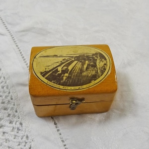 May include: A small, rectangular wooden box with a hinged lid and a metal clasp. The lid features an oval image of a pier and railway station. The box is light brown with a glossy finish, and the image has a sepia tone.