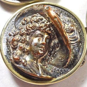 Peut inclure: Un bouton métallique circulaire présentant un relief tridimensionnel d'une personne aux cheveux bouclés portant un chapeau à larges bords. Le bouton possède un bord doré poli et une finition sombre et vieillie sur le portrait central.