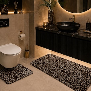 May include: A two-piece bathroom rug set featuring a leopard print pattern in shades of brown, black, and tan. The set includes one rectangular rug and one smaller square rug with a cutout designed to fit around the base of a wall-mounted toilet.