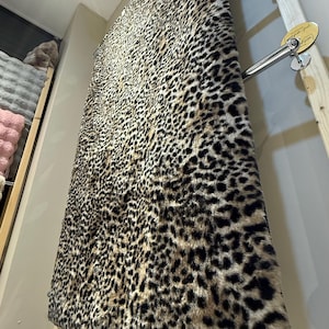 May include: A large, faux fur rug with a leopard print pattern. The rug is primarily tan with black spots and is displayed vertically. The rug is hanging on a metal rack, showcasing its texture and design.