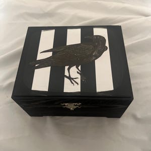 May include: A black wooden box with a hinged lid and a silver clasp. The top of the box features a black and white striped design with a detailed illustration of a crow. The crow is dark brown and has its wings folded.