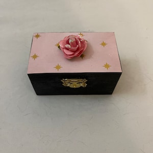 May include: A small, black wooden box with a pink lid decorated with gold stars and a pink flower. The box has a gold latch.