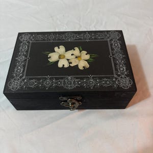 May include: A rectangular black wooden box featuring a white floral design of two dogwood blossoms on the lid. The lid is decorated with a white intricate filigree border. A metallic latch is centered on the front side of the box.