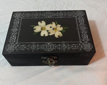 May include: A rectangular black wooden box featuring a white floral design of two dogwood blossoms on the lid. The lid is decorated with a white intricate filigree border. A metallic latch is centered on the front side of the box.