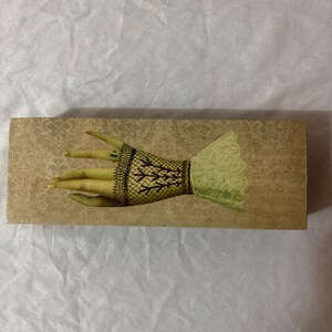 May include: A rectangular wooden box featuring a beige damask pattern background. The top surface displays a graphic illustration of a hand wearing a dark brown fishnet glove with a green ring on one finger and a light green ruffled cuff.