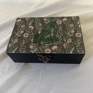 May include: A black wooden box with a decorative lid featuring a green frog illustration. The lid has a floral pattern with pink and white flowers on a dark background. The box has a metal clasp.