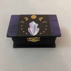 May include: A small wooden box with a purple lid and a black base. The lid has a decorative design featuring a crystal, moons, and stars in gold and pink.