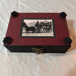May include: A rectangular wooden box with a dark maroon lid and black sides. The lid features a black and white vintage photograph of a horse-drawn carriage with the text J. HISLOP UNDERTAKER. Four black rose ornaments are attached to the corners.