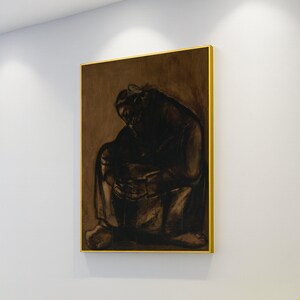 Puede incluir: Una obra de arte enmarcada representa una figura sentada en tonos oscuros sobre un fondo marrón. La figura está encorvada, con la cabeza gacha. La obra de arte está encerrada en un marco dorado, creando un contraste con los tonos más oscuros de la imagen.