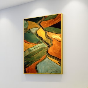 May include: Abstract landscape painting in a gold frame. The artwork presents a vibrant mix of colours, including shades of green, gold, orange, and blue, depicting a stylized aerial view of fields and waterways. The painting is mounted on a white wall.