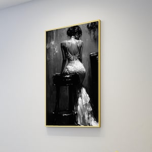 May include: Black and white framed artwork depicting a woman in a formal gown. The woman is seated on a stool, her back to the viewer. The dress has a low back and a long train. The artwork is in a gold frame.