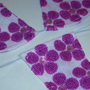 May include: A white bikini top and bottom set featuring a vibrant purple sequin floral pattern. The fabric is covered in circular purple sequins arranged in flower shapes, connected by thin, light-colored elastic strings for adjustable ties.