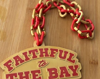 May include: A gold and red pendant with the text "FAITHFUL to THE BAY" and a matching chain. The pendant and chain are made of a shiny material and are displayed on a wooden surface.