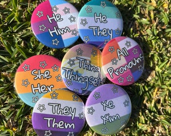 May include: A collection of seven round pronoun buttons with colorful backgrounds and star accents. Each button displays a different set of pronouns, including "He/Him", "He/They", "She/Her", "They/Them", "Xe/Xim", "All Pronouns", and "Thing/Thingself".