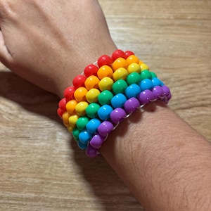 May include: A rainbow-colored beaded bracelet. The bracelet features rows of red, orange, yellow, green, blue, and purple beads. The beads are strung together on a clear elastic band, creating a vibrant and eye-catching accessory.