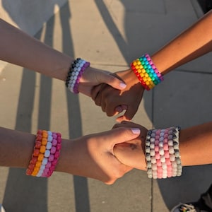 Puede incluir: Cuatro pulseras de cuentas de colores en una variedad de colores, incluyendo arcoíris, rosa, naranja y blanco. Las pulseras se usan en las muñecas, con las manos entrelazadas. Las pulseras están hechas de pequeñas cuentas redondas.