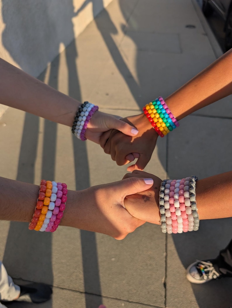 May include: Four colorful beaded bracelets in a variety of colors, including rainbow, pink, orange, and white. The bracelets are worn on wrists, with hands clasped together. The bracelets are made of small, round beads.