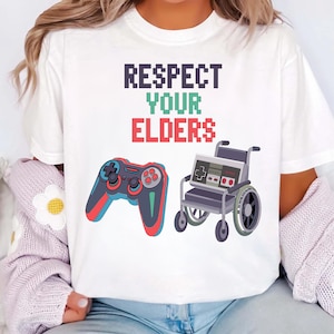 May include: White t-shirt with the text "RESPECT YOUR ELDERS" in pixelated font. The design includes a video game controller and a wheelchair, both in a cartoon style. The controller is red, blue, and black.