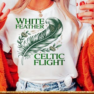 Peut inclure: T-shirt blanc avec un motif de plume celtique verte. Le texte sur le t-shirt indique "WHITE FEATHER" au-dessus de la plume et "CELTIC FLIGHT" en dessous. La plume est ornée de motifs de nœuds celtiques et d'éléments floraux.