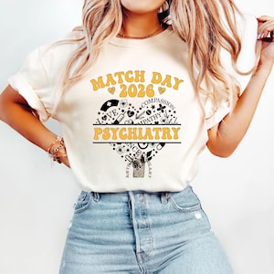 Peut inclure: T-shirt crème avec l'inscription "MATCH DAY 2026 PSYCHIATRY" dans une police rétro. Le motif comprend un cœur avec des icônes médicales et les mots "LOVE" et "COMPASSION".
