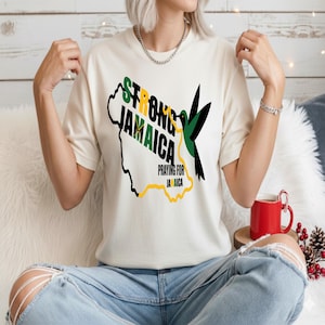 Könnte beinhalten: Cremefarbenes T-Shirt mit dem Text "STRONG JAMAICA" in Grün und Schwarz, einer Kartenumriss und einer Kolibri-Grafik. Das Shirt enthält auch den Satz "PRAYING FOR JAMAICA". Im Hintergrund ist eine rote Tasse zu sehen.