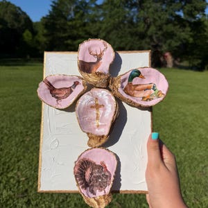 May include: A decorative cross made of oyster shells, each with a different animal illustration. The shells are set on a white canvas with gold trim. The animals include a deer, duck, pheasant, and turkey. A gold cross is in the center.