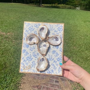 May include: A decorative wall art piece featuring five oyster shells arranged in a cross shape. The shells are set against a blue and white floral patterned background with a gold border. The initials "SBS" are visible in the lower left corner.