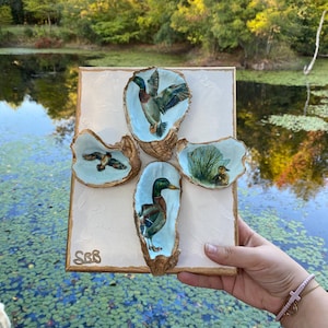 May include: A decorative wall art piece featuring four painted oyster shells with duck illustrations. The artwork is mounted on a textured, off-white canvas with a gold-colored frame. The initials "SGB" are visible in the lower left corner.