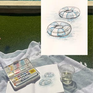 May include: Watercolor painting of two blue and white striped pool floats on water. A watercolor palette, sketchbook with a drawing of the floats, and a clear cup of liquid are on a towel.