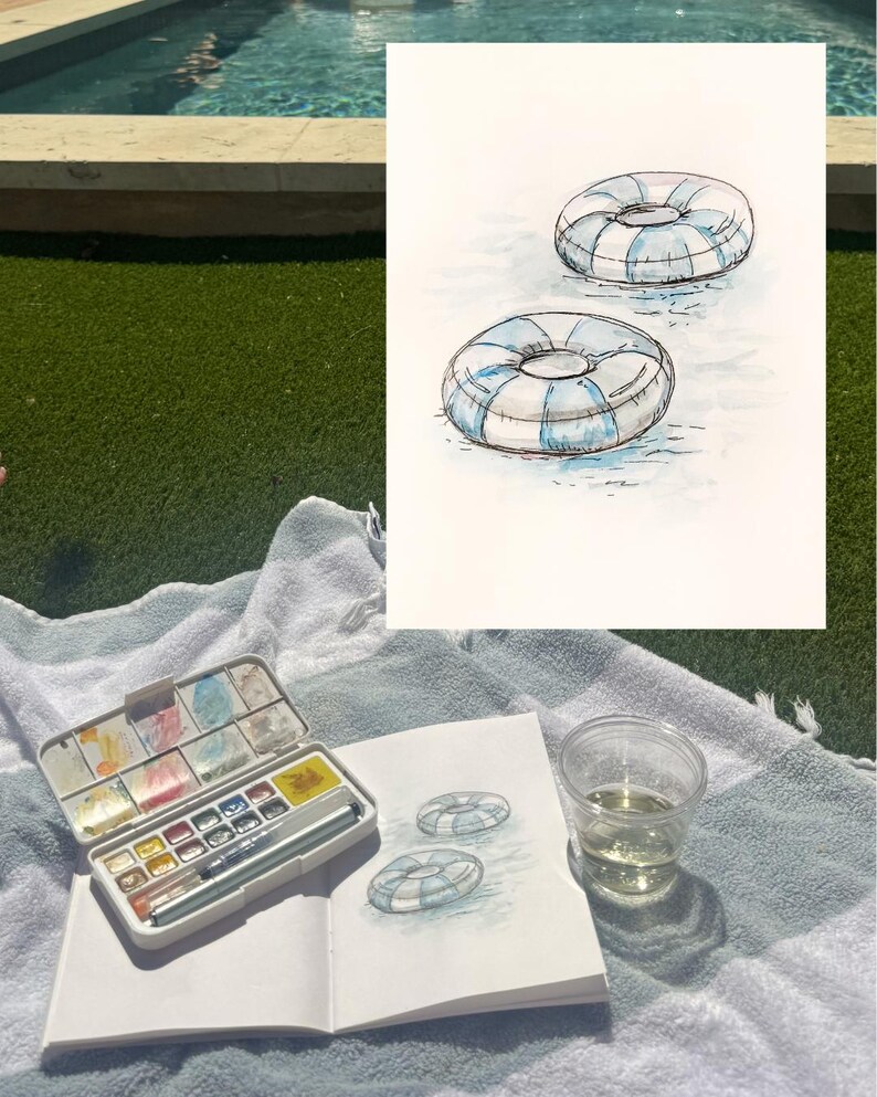 May include: Watercolor painting of two blue and white striped pool floats on water. A watercolor palette, sketchbook with a drawing of the floats, and a clear cup of liquid are on a towel.