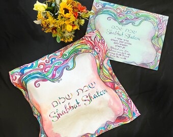 Può includere: Un tovaglietta di stoffa bianca con un design astratto colorato e il testo ebraico "Shabbat Shalom" in lettere nere. La tovaglietta è su una superficie nera.