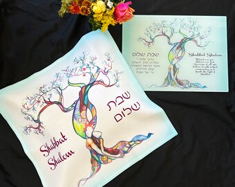 Può includere: Un panno da Shabbat bianco con un design a forma di albero della vita colorato. L'albero è intrecciato con due figure, e le parole ebraiche "Shabbat Shalom" sono scritte in bordeaux. Il panno è su una superficie nera.